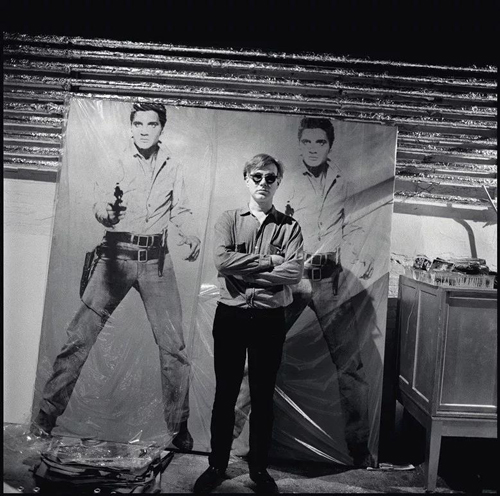 艺术品：© 2018The Andy Warhol Foundation for the Visual Arts, Inc. / Licensed by Artists Rights Society (ARS)，照片：© Bruce Davidson/Magnum Photos。 安迪‧沃荷与《双面猫王》 于工作室的合影，约1964年